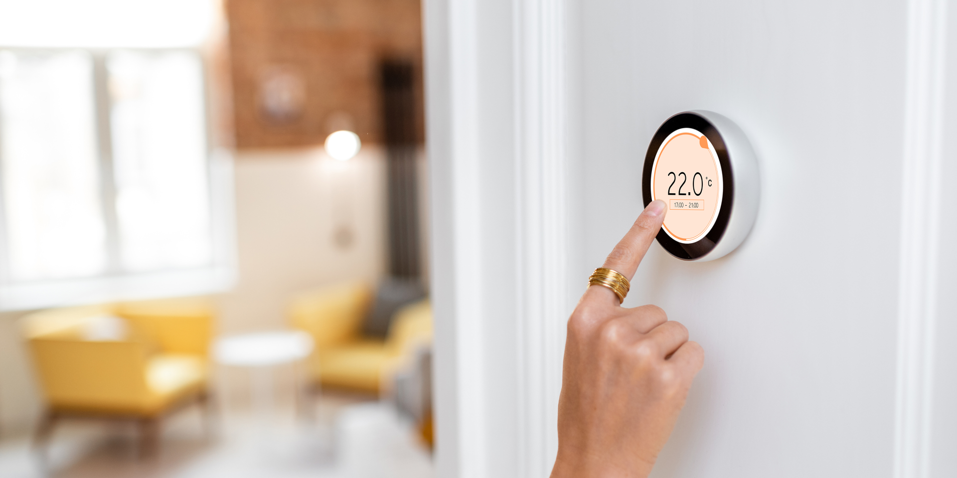 A person's hand touches a smart thermostat displaying 72 degrees. The device is mounted on a white wall in a living room.