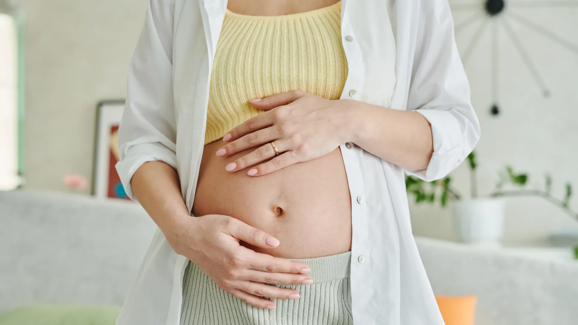 Pregnant woman gently holding her belly indoors, wearing a yellow top and open white shirt
