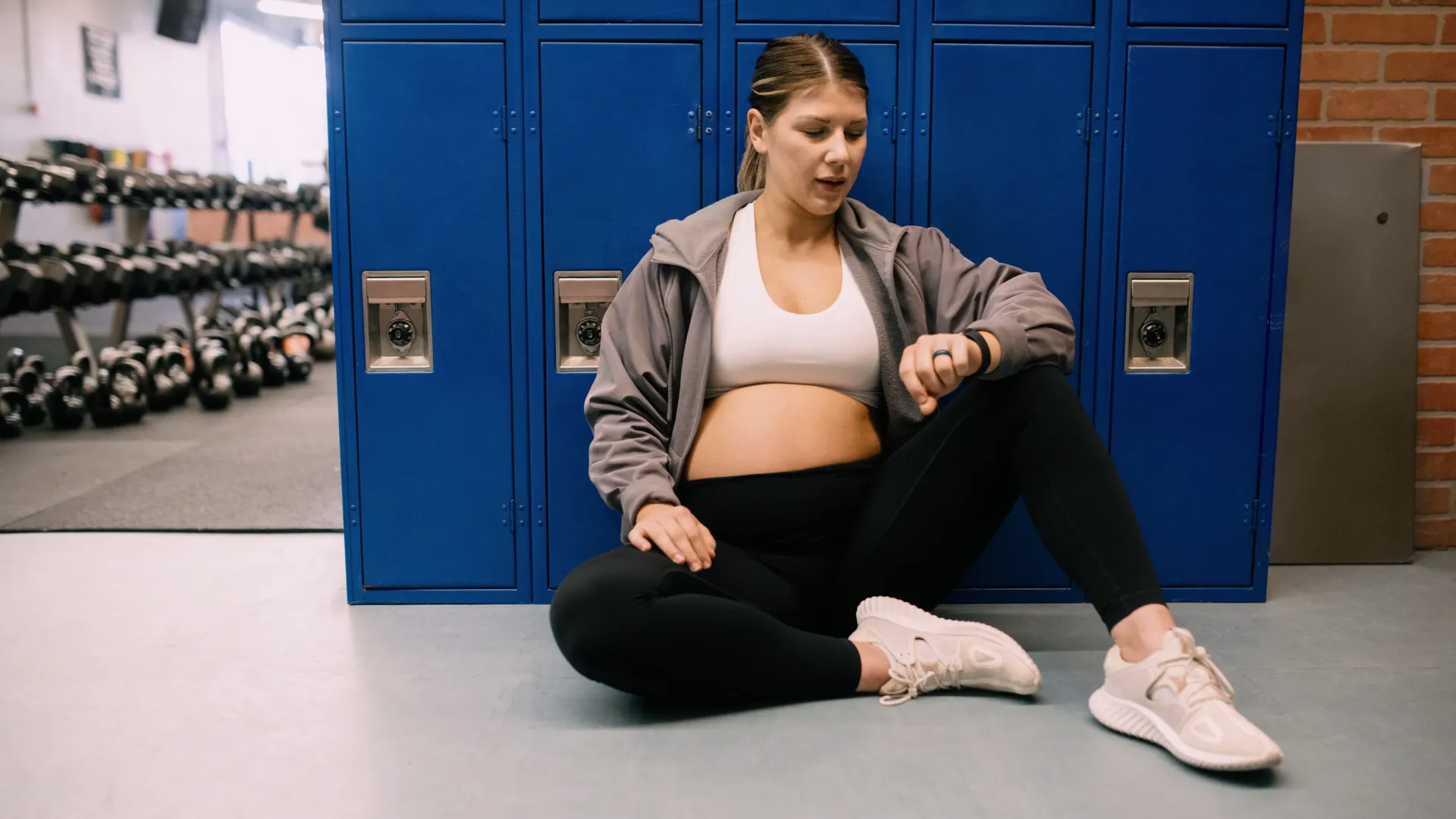 Pregnant woman sitting on a gym floor checking her fitness watch after a workout