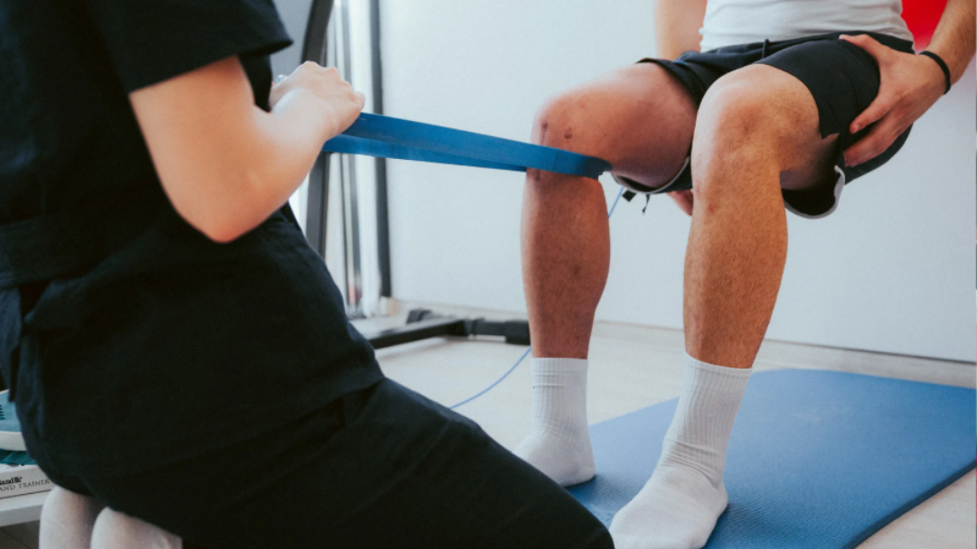 Coach guiding knee rehabilitation with a resistance band as the client performs controlled movement on a blue mat.
