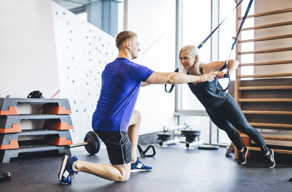 A fitness coach in blue instructing a woman using suspension training straps in a modern gym