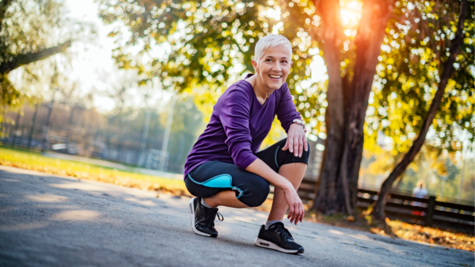 Smiling active woman in athletic wear kneeling in a sunny park after a workout.