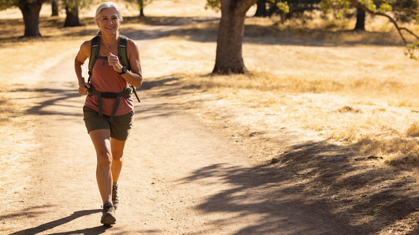 A mature woman finishing a sunlit trail hike with energy to spare, everyday capacity and independence.