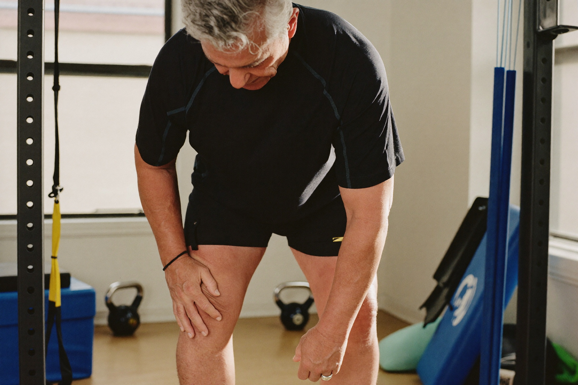 A mature adult pausing beside a squat rack, hand on a sore knee, deciding whether to train.