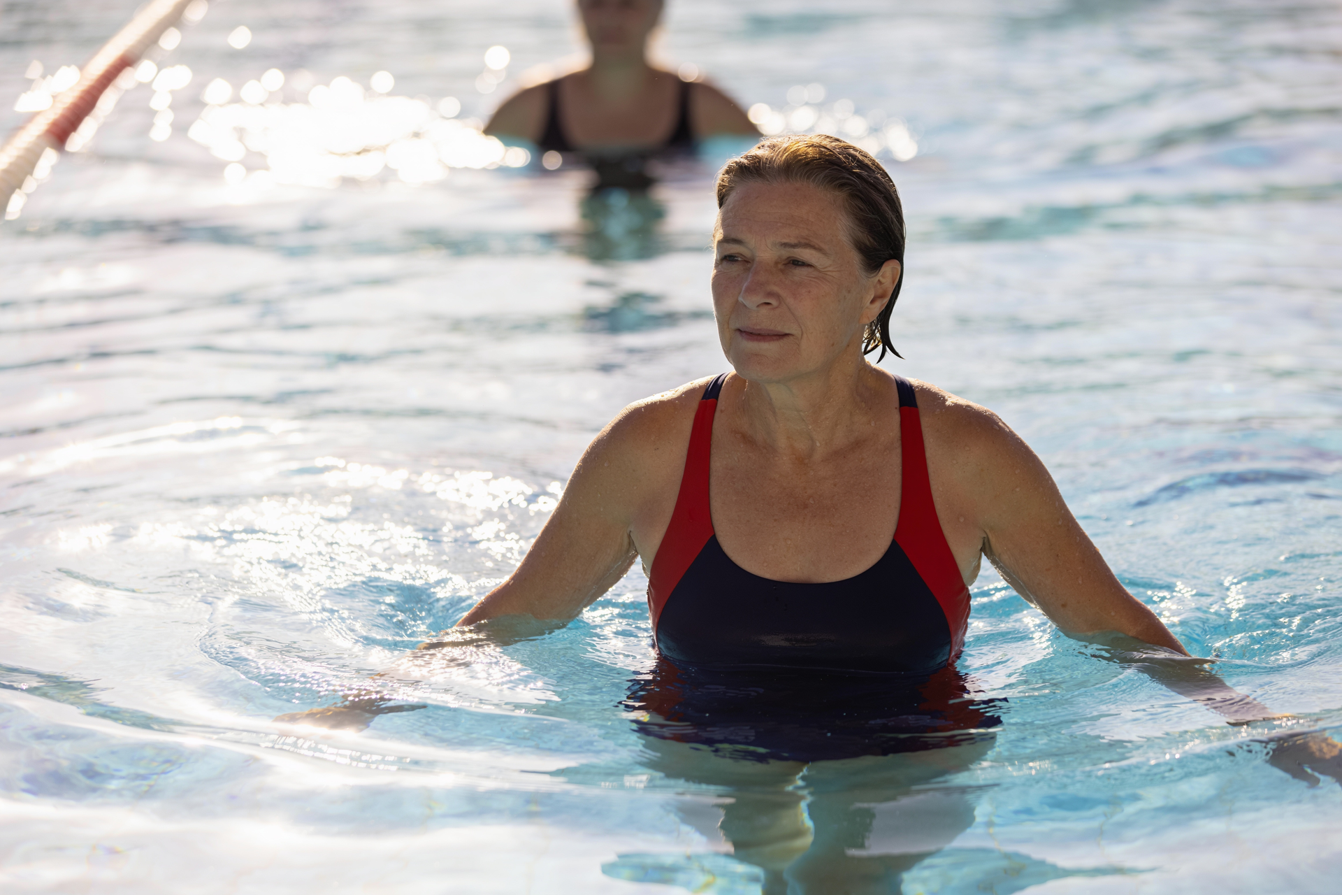 An older adult water-walks in a sunlit pool, exercising while staying cool, with another swimmer blurred behind.