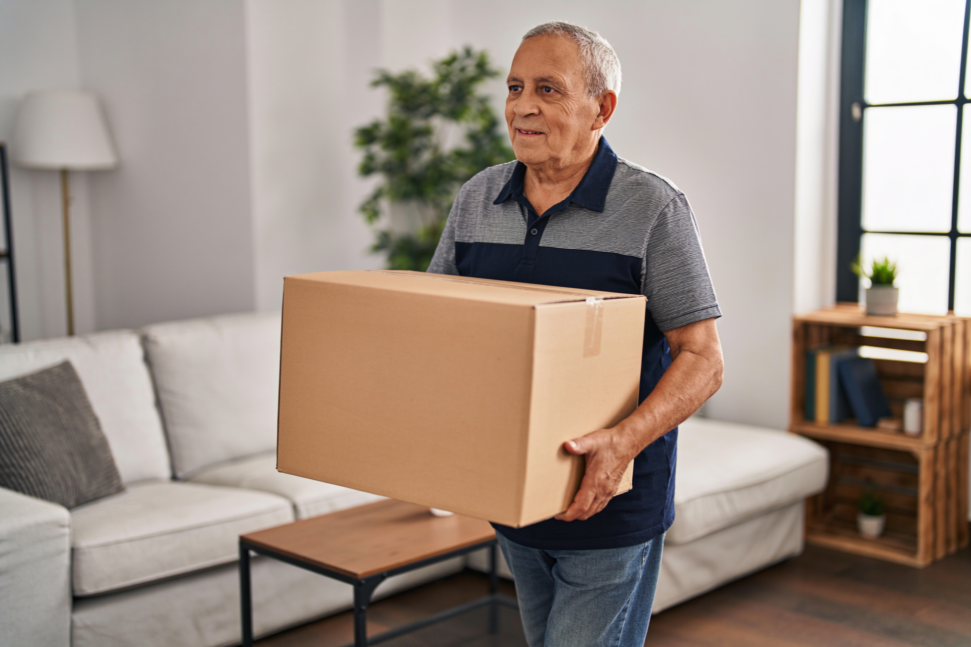 A man in his early 60s stands holding a cardboard box at waist height, about to carry it across a room with controlled posture.