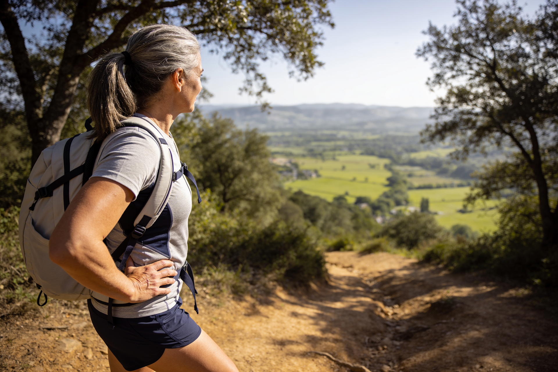 A woman in her fifties hikes a sunlit trail with a daypack, pausing at a valley view.