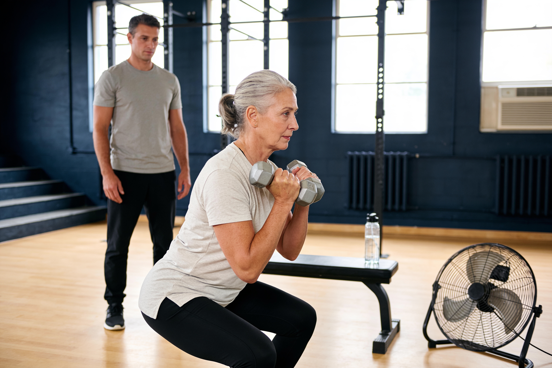 A mature woman performs a controlled goblet squat indoors with a fan and a water bottle nearby.