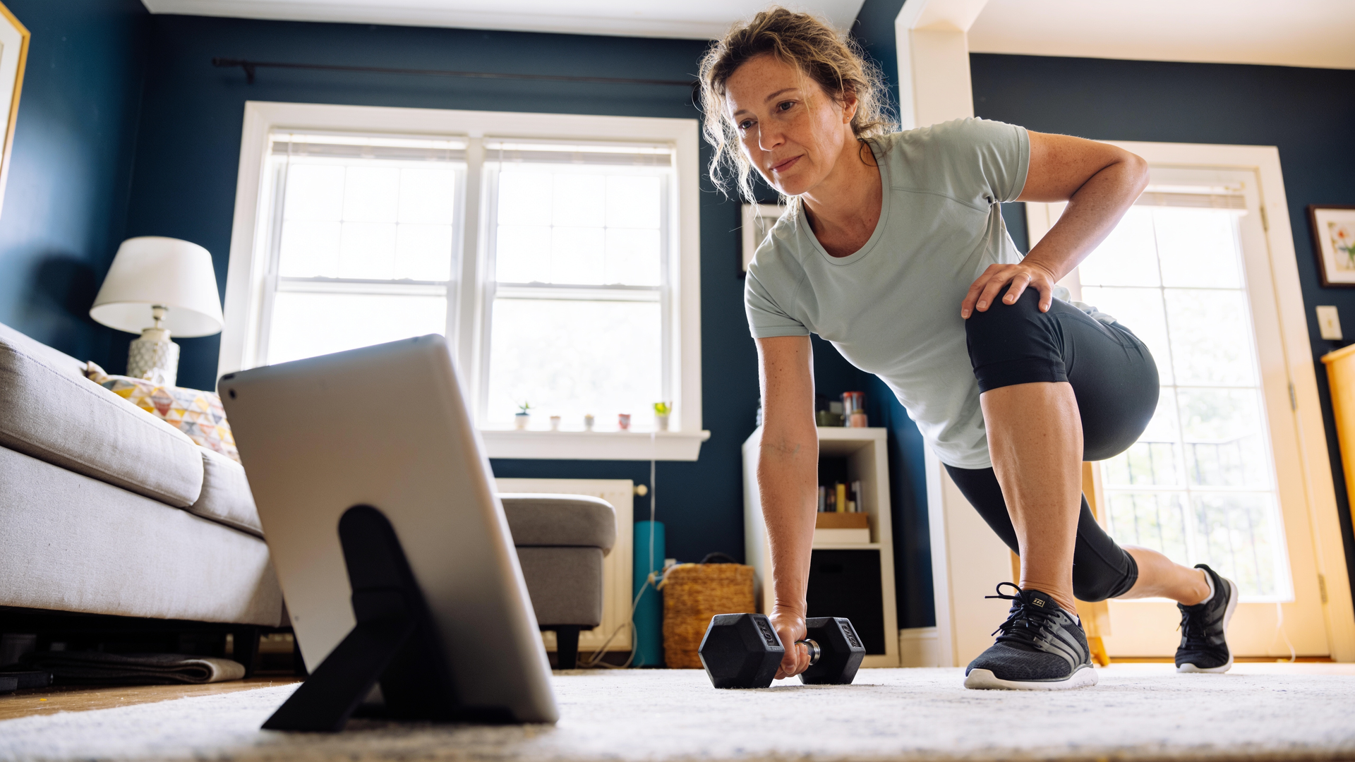 A woman does a forward lunge in her living room while following a tablet propped in front of her.