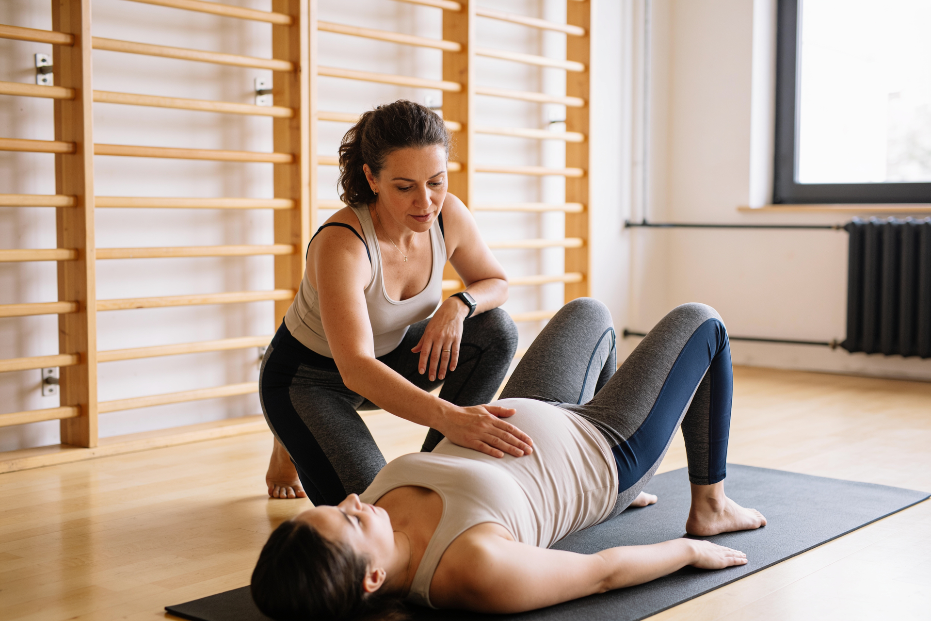 A coach kneels beside a postpartum client lying on her back and cues a slow heel slide, one hand guiding her knee as she keeps her pelvis still.