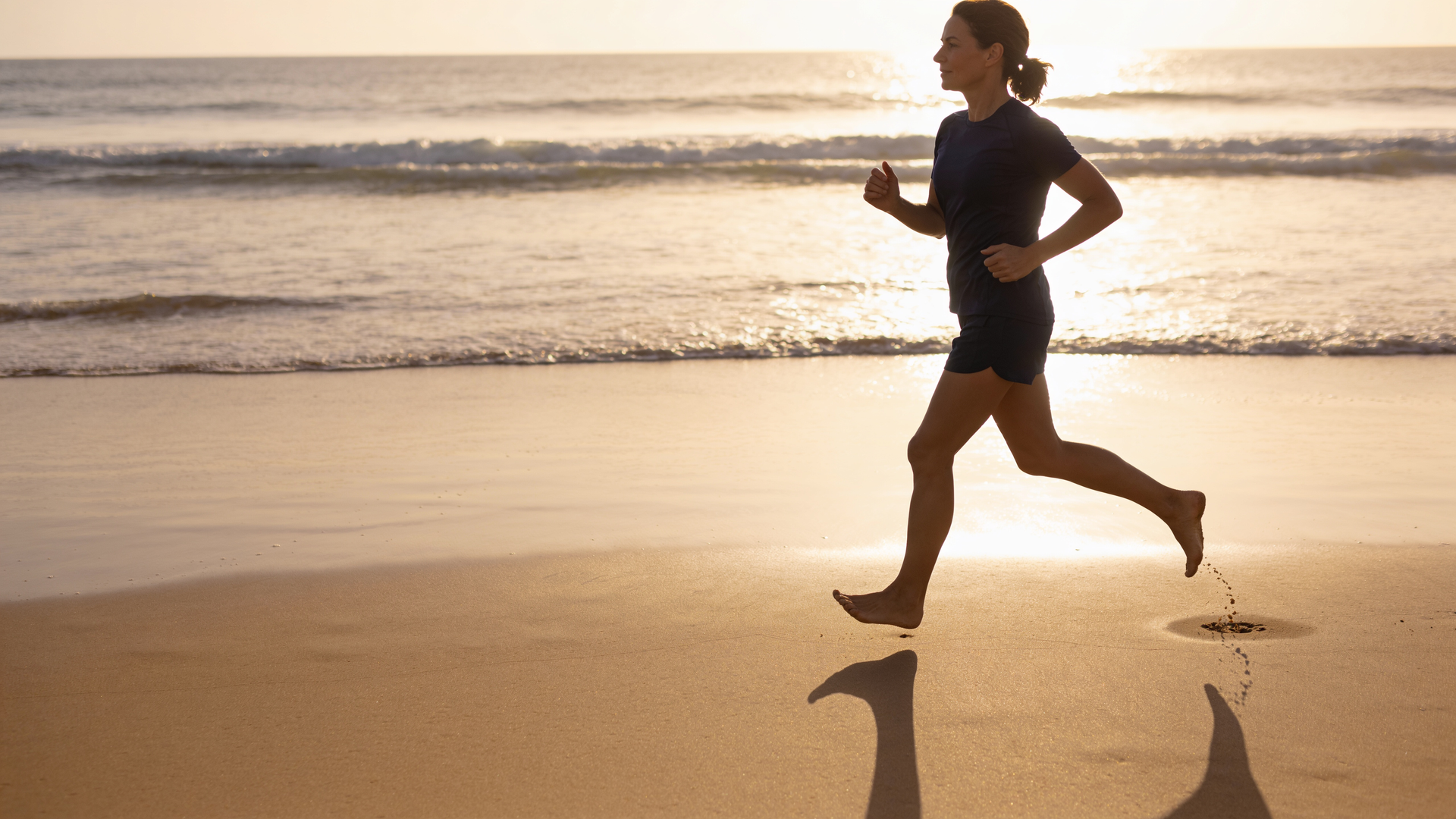 A woman power-walks along firm wet sand at the waterline in soft morning light.