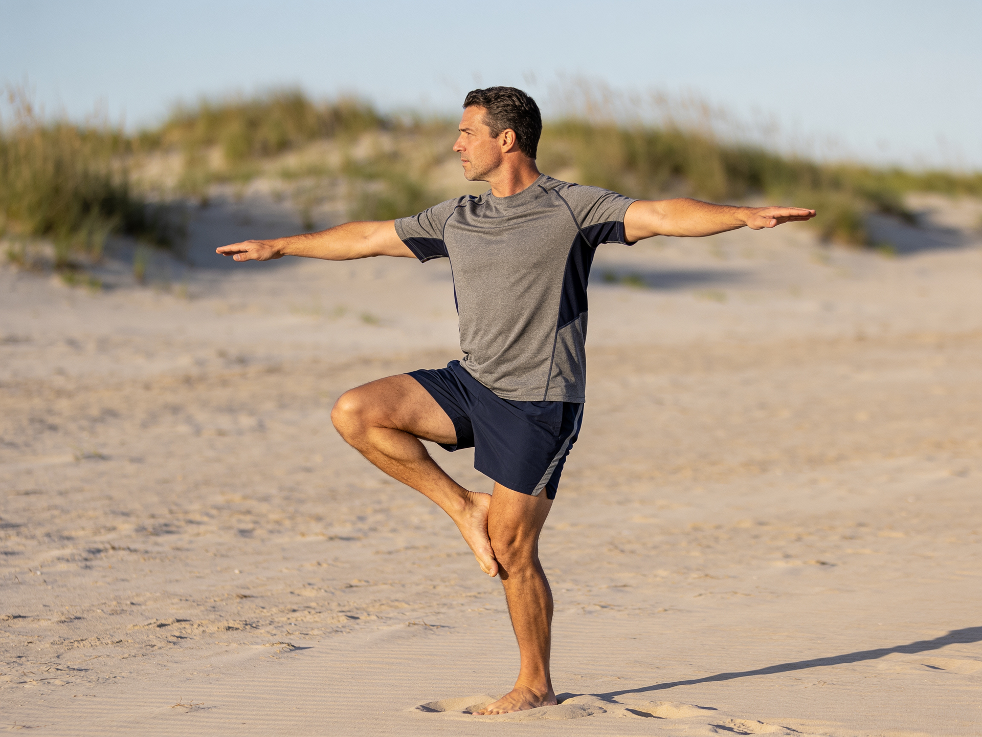 A man holds a single-leg balance on soft dry sand, arms out to steady himself.