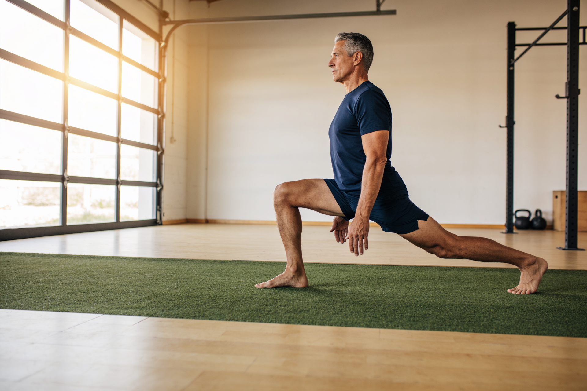 An adult holds a deep, controlled long-stride forward lunge in a bright, airy setting, showing earned full range of motion.