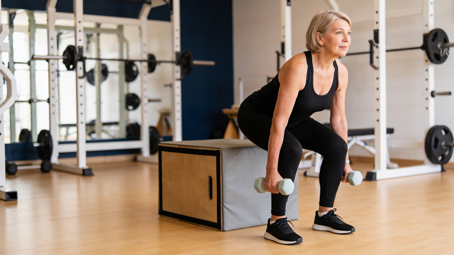 Woman in her fifties doing a controlled supported squat to a box with light dumbbells.