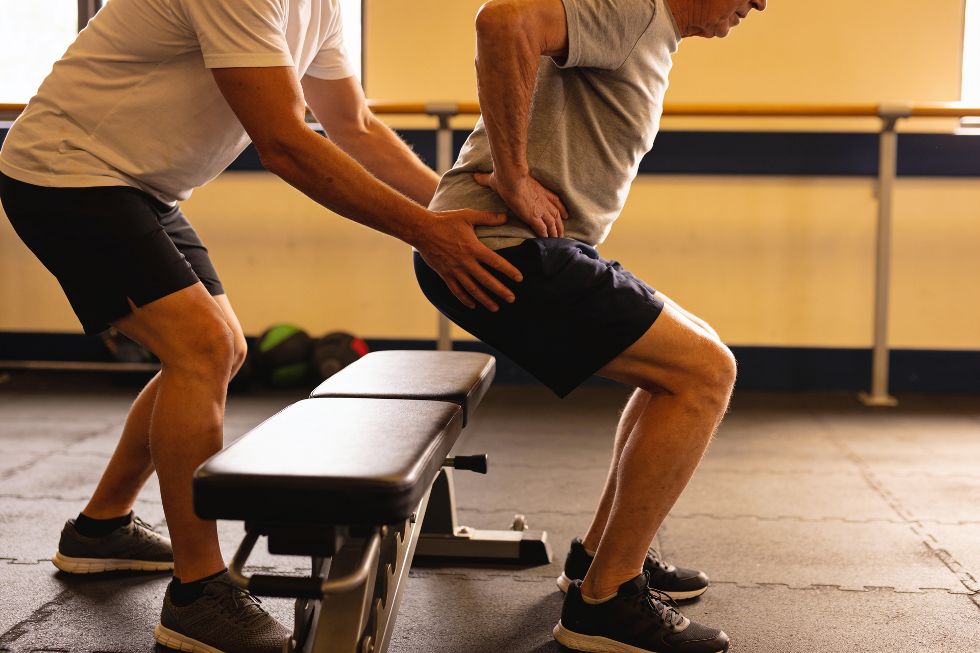 A coach guiding an older adult through a sit-to-stand exercise to a bench, lightly cueing the hips.