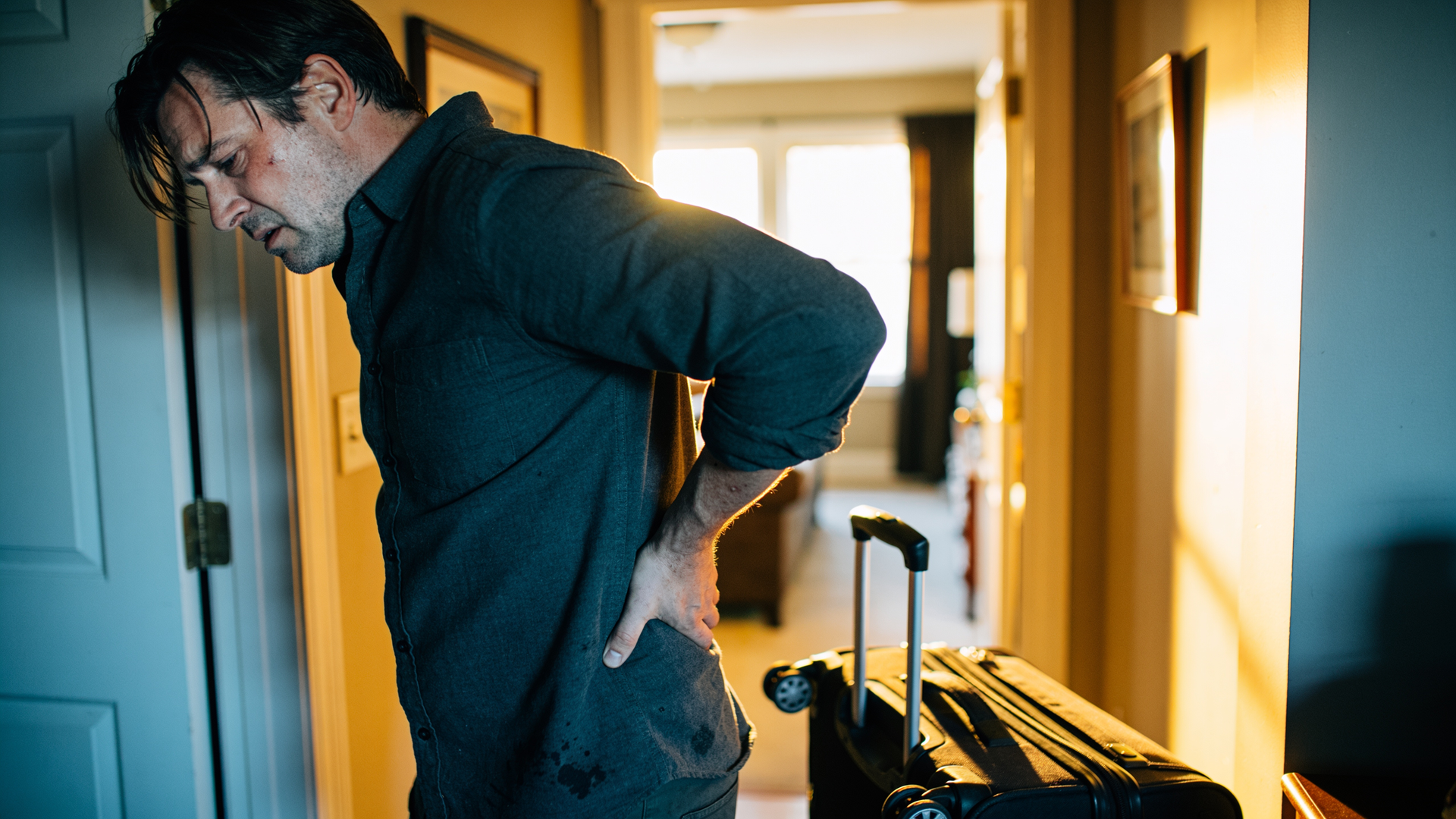 An adult just home from travel easing a tight lower back, jet-lagged and stiff, with a suitcase near