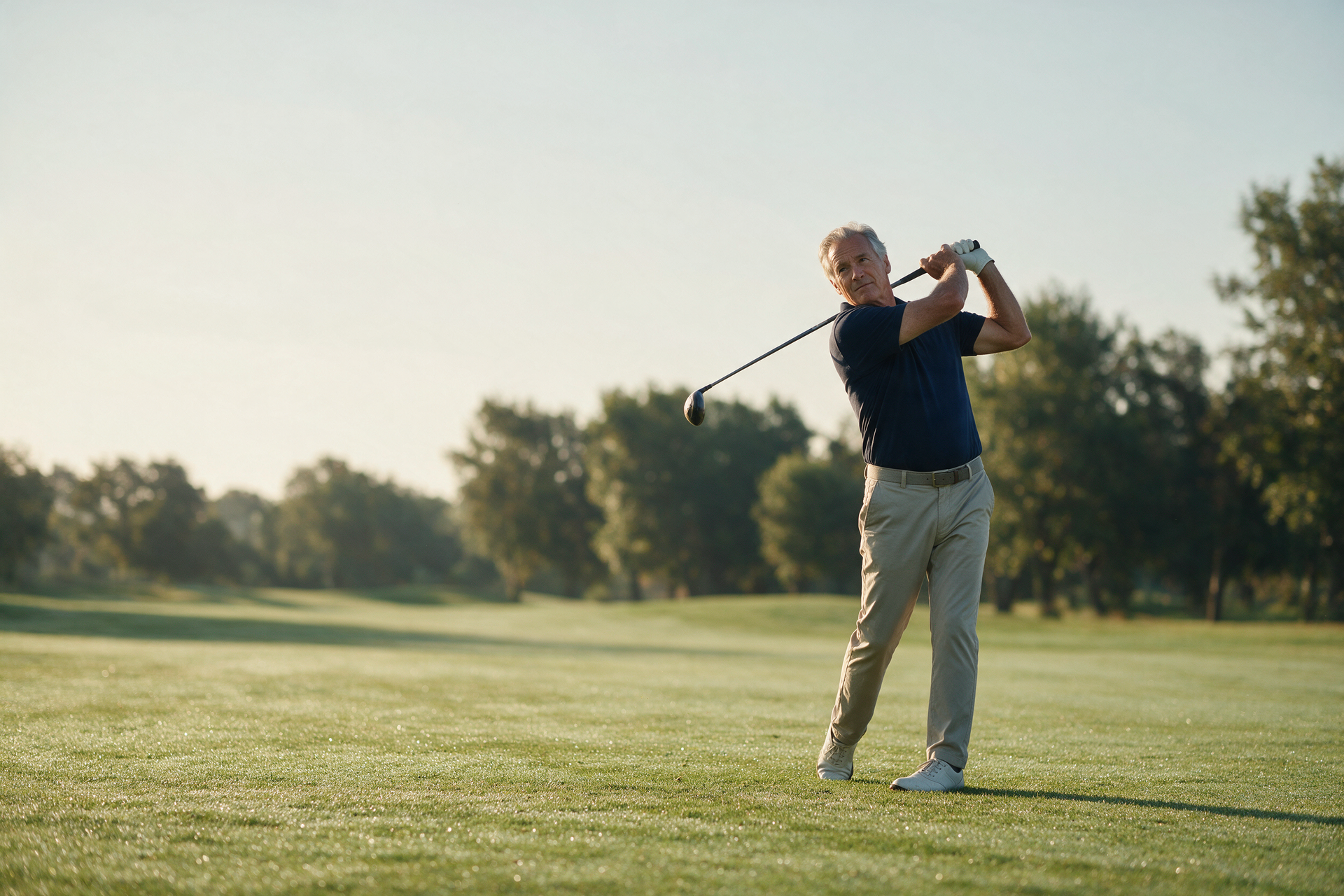 A mature golfer finishes a full swing on a quiet fairway in soft morning light.