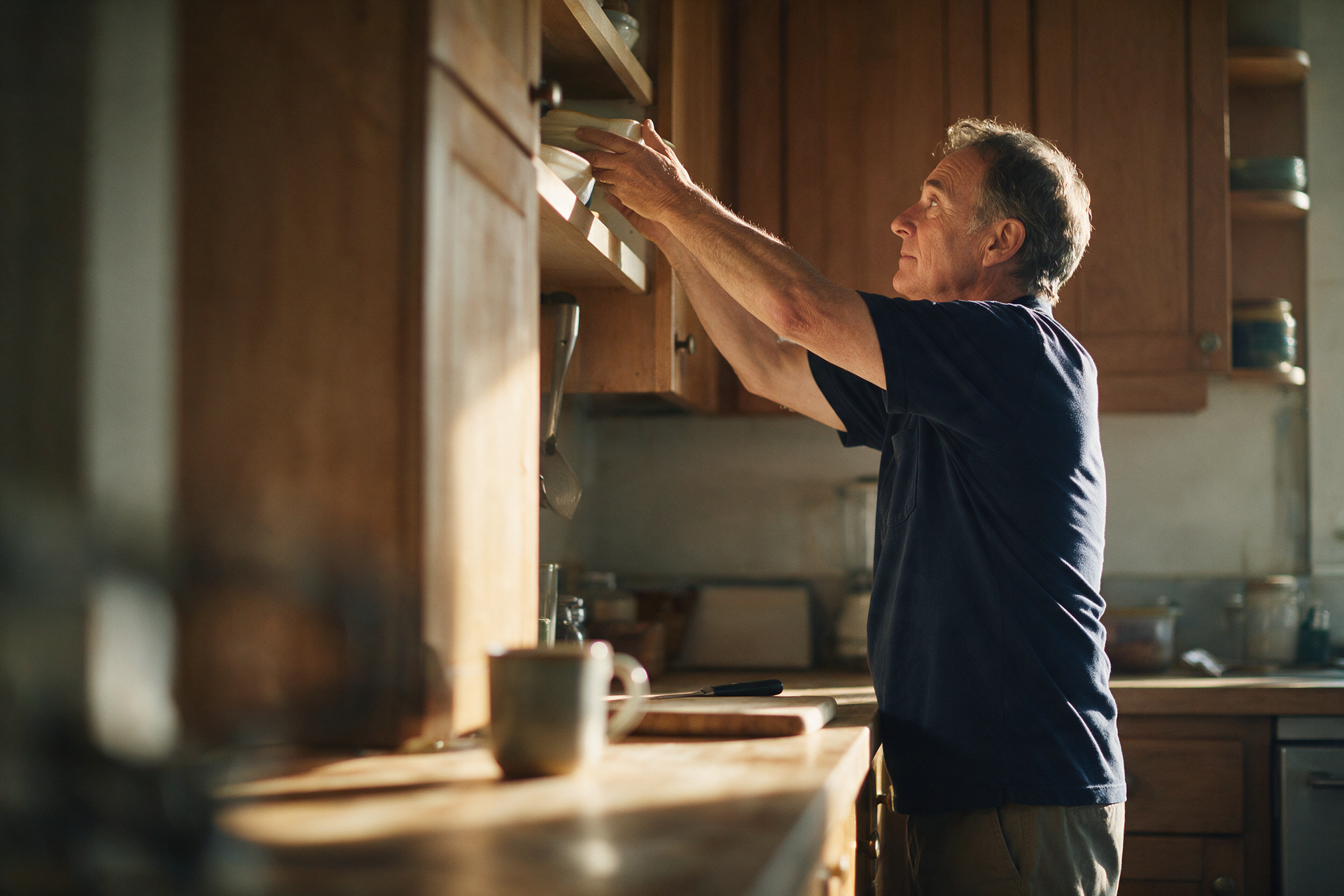 Mature man reaches to place a dish on a high kitchen shelf in soft morning light, a hint of caution