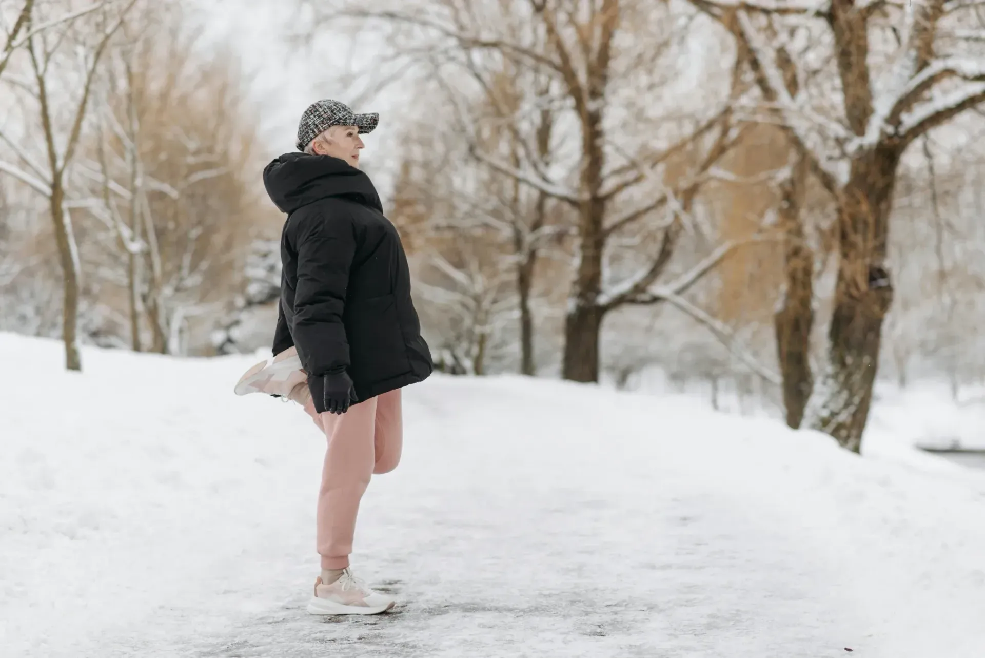Person in winter clothing standing on a snowy path stretching their leg behind them