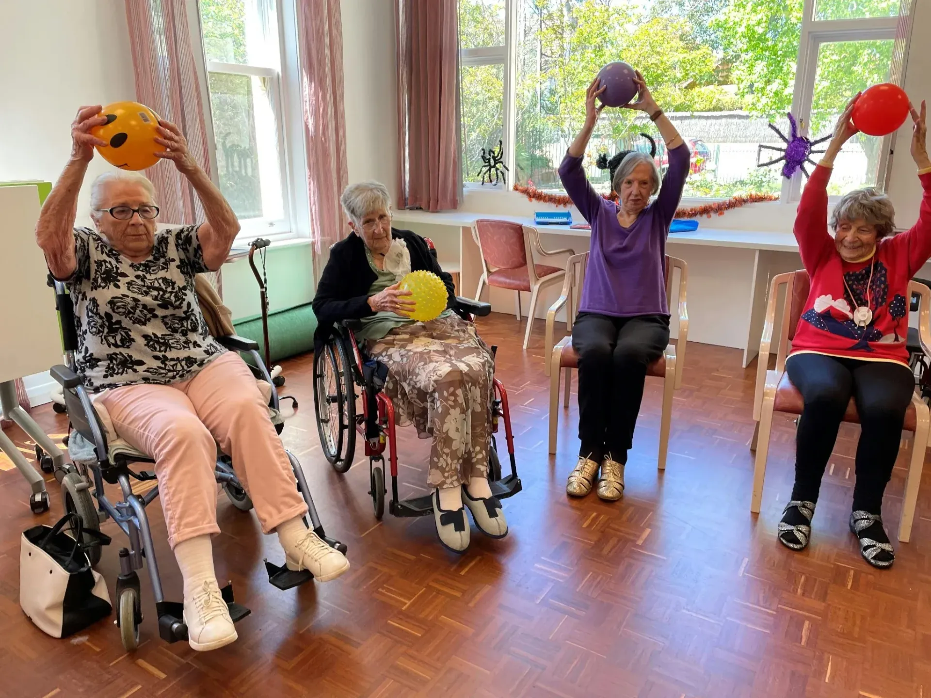 Group of older adults seated indoors raising exercise balls overhead, engaging in gentle fitness activity