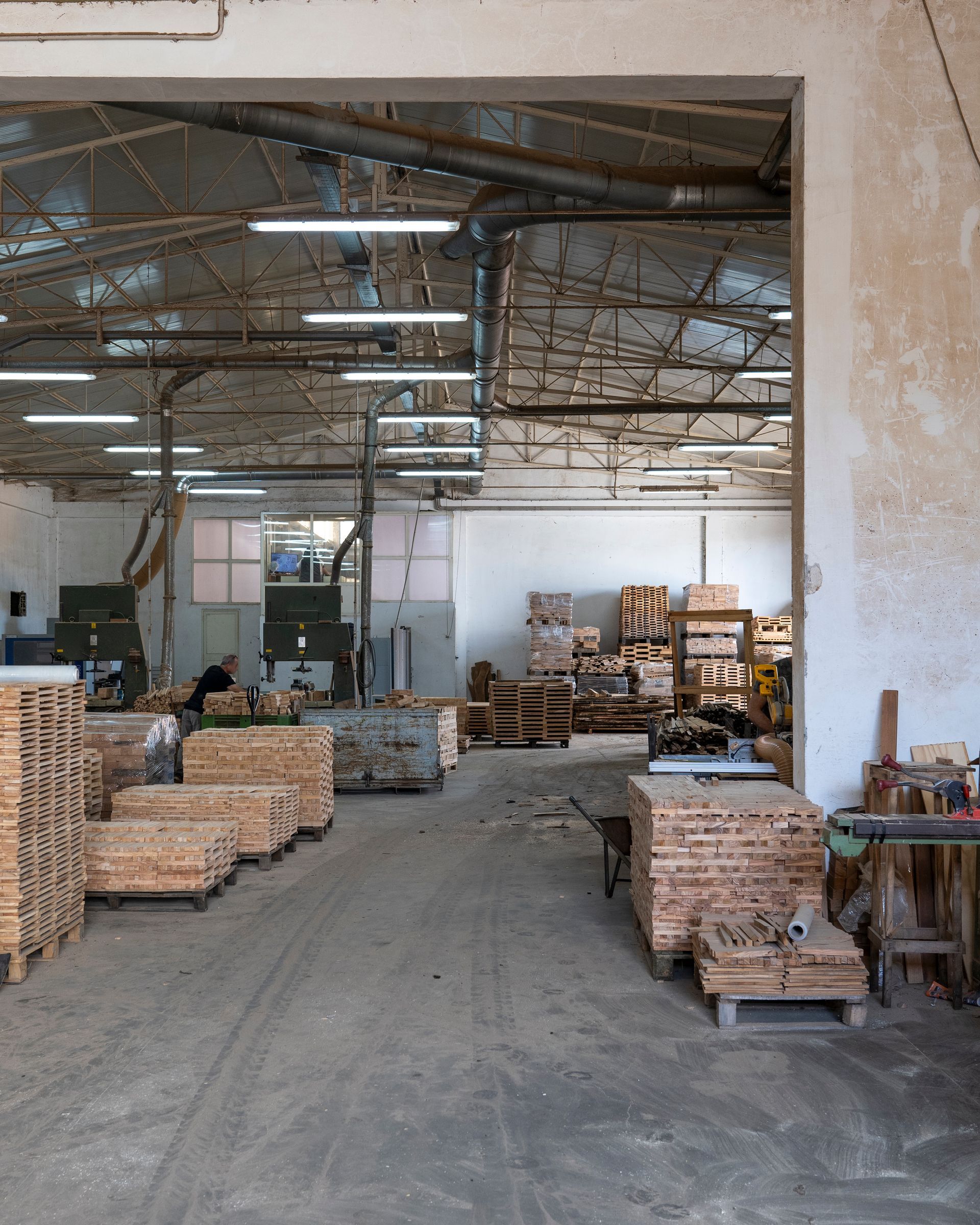Interno di un magazzino con pile di pallet e casse di legno tagliati sotto un alto soffitto metallico.