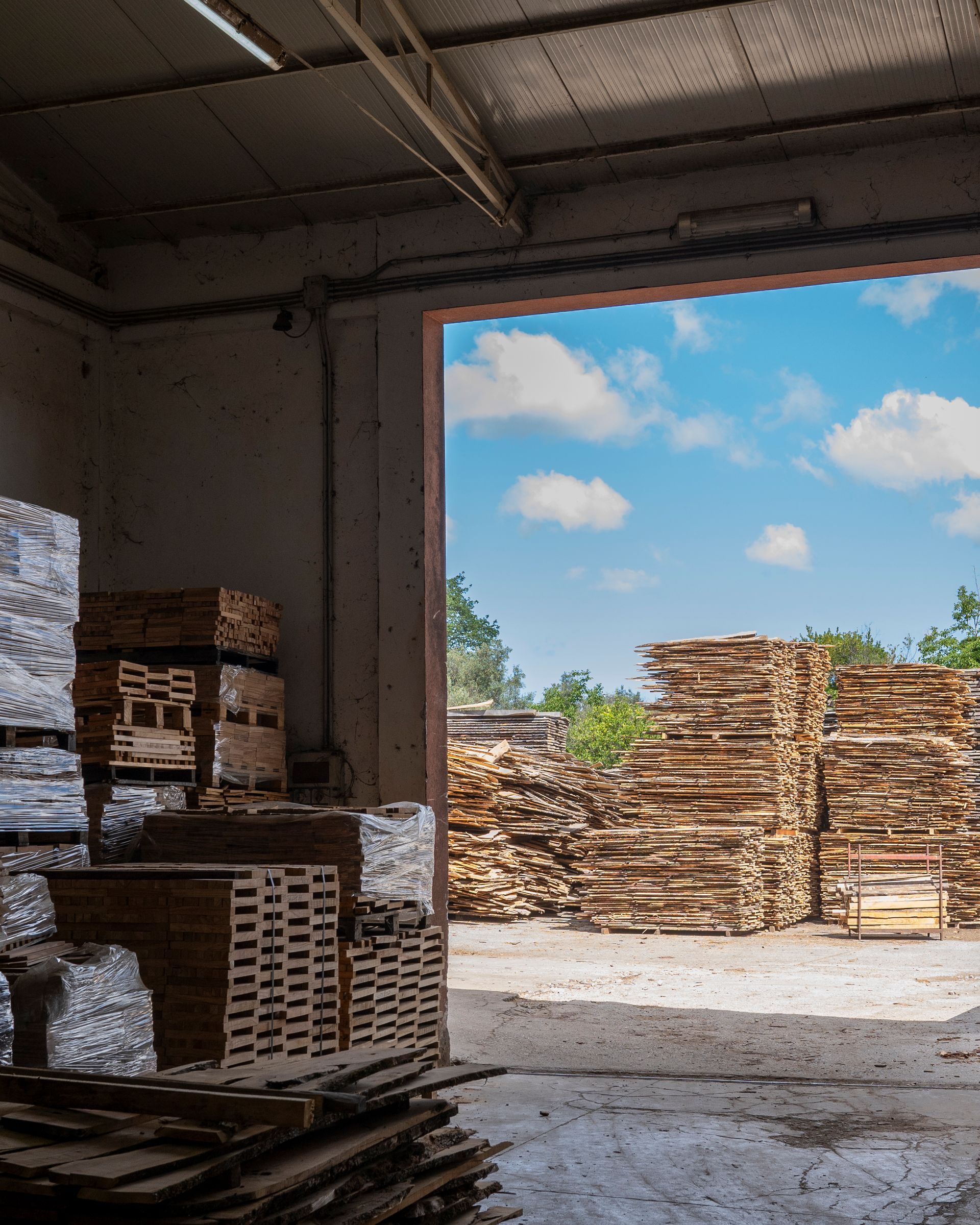 Pile di pallet di legno stoccati in un magazzino, con il cielo azzurro visibile attraverso la porta aperta.