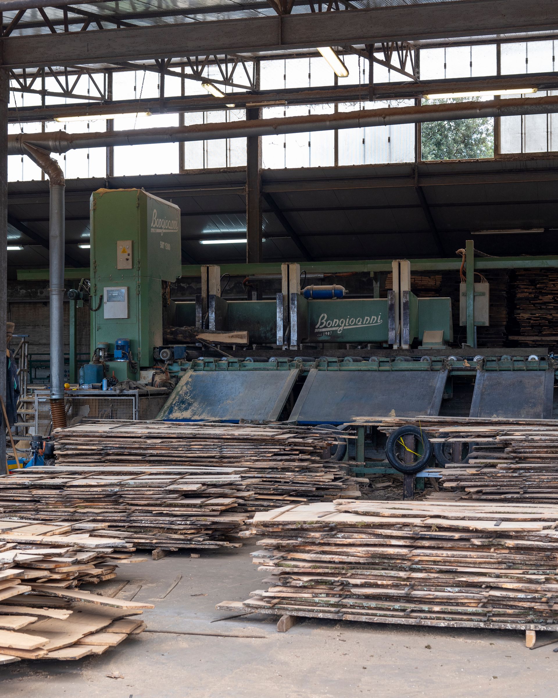 Interno di una segheria industriale con cataste di legname tagliato e una grande macchina da taglio verde.