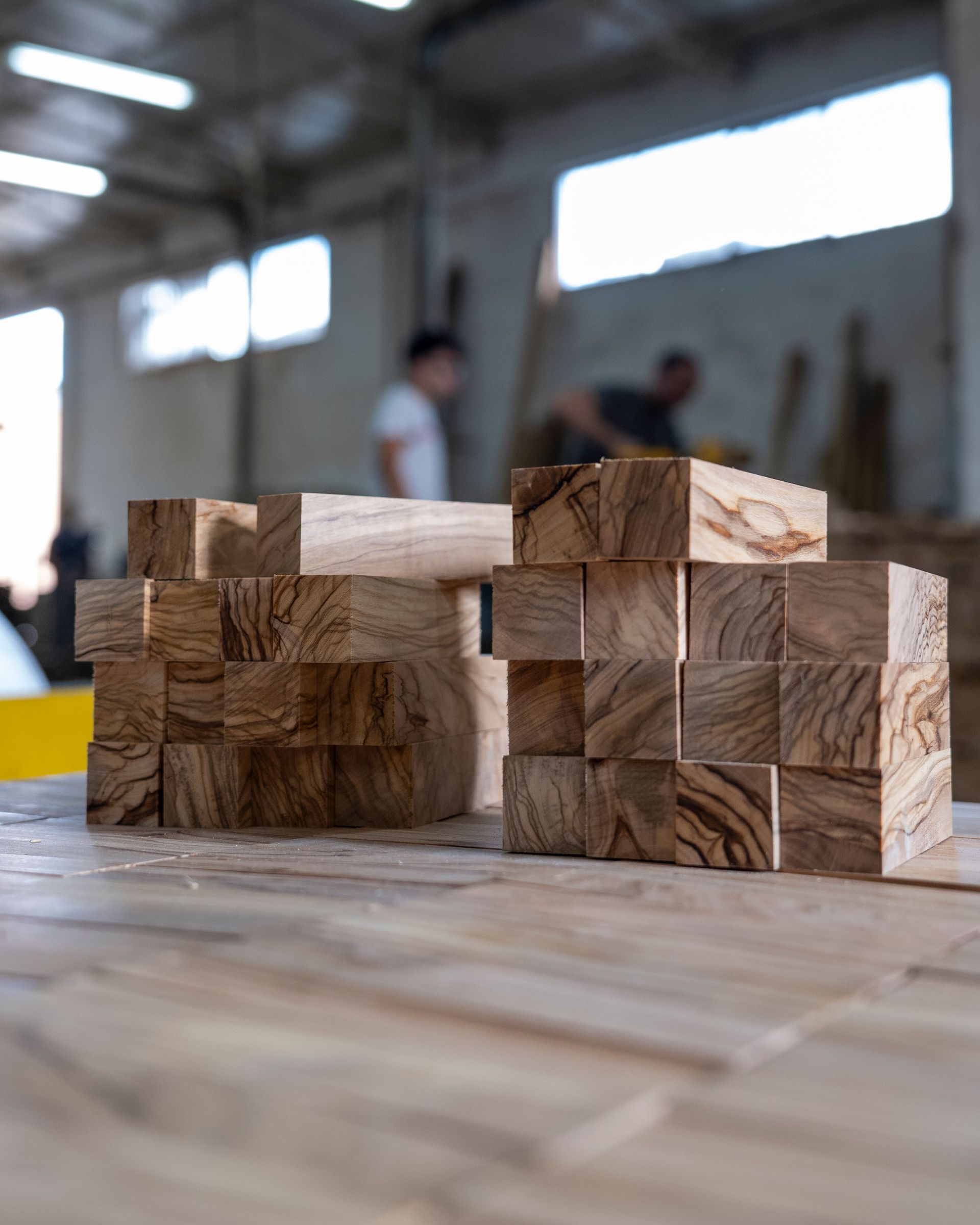 Blocchi di legno impilati su un banco da lavoro in un'officina, con persone sfocate sullo sfondo.