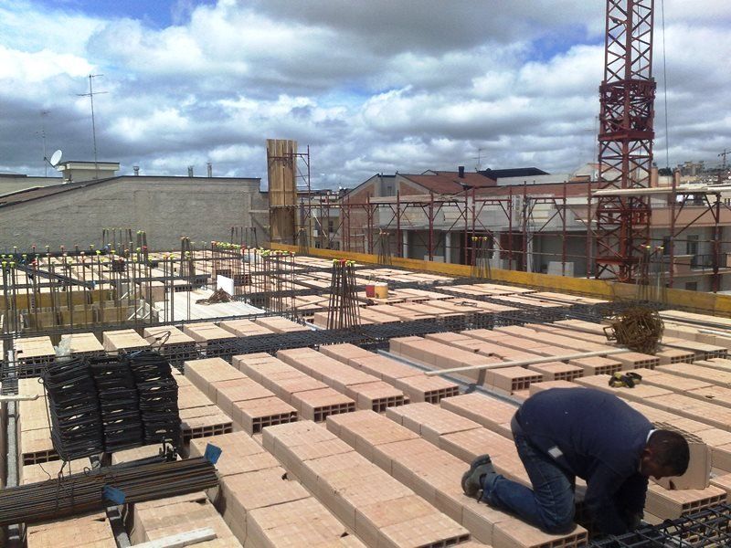 Un operaio edile inginocchiato su un'impalcatura di mattoni di terracotta e barre d'acciaio in un cantiere attivo.