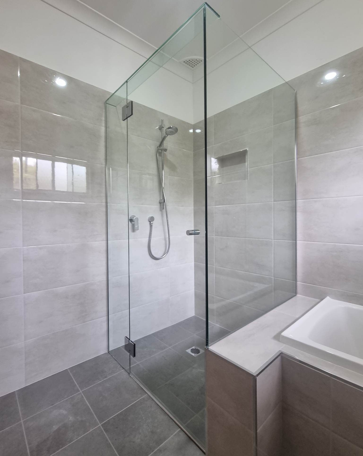 Glass shower enclosure with chrome fixtures in a modern bathroom with gray tile and a built-in ledge — K & J Glass in Valla Beach, NSW