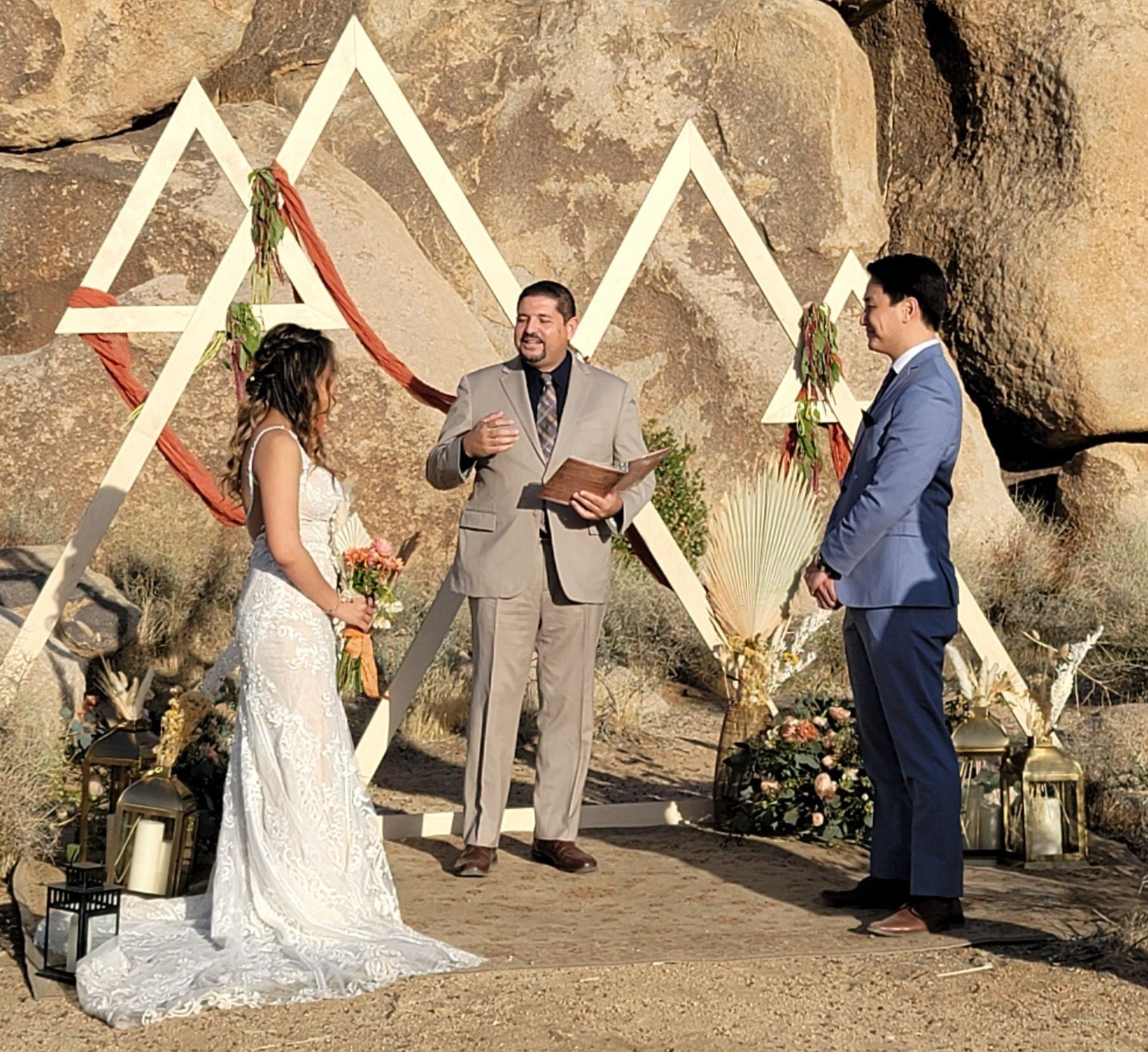 Wedding ceremony outdoors: couple, officiant, triangle arch, against rock formations.