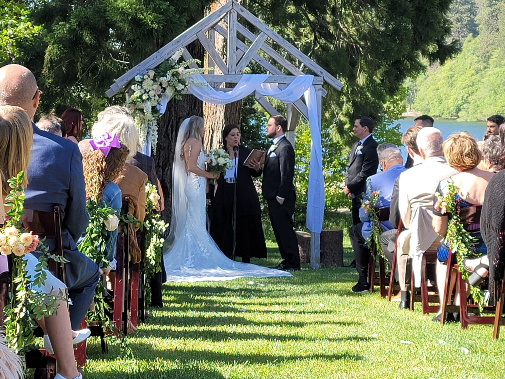 Wedding ceremony outdoors with couple under decorated arbor, guests seated on either side.