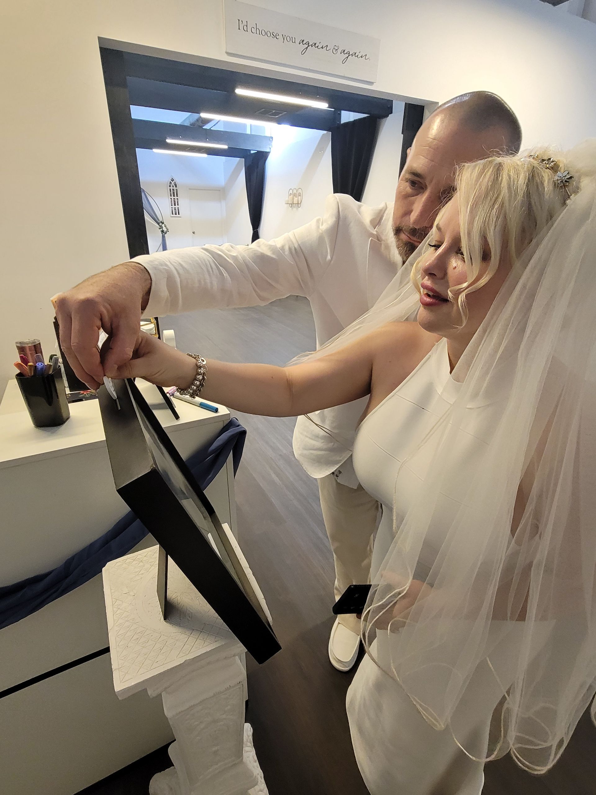 Bride and groom sign a framed photo; bride wears veil, groom in white shirt, inside.