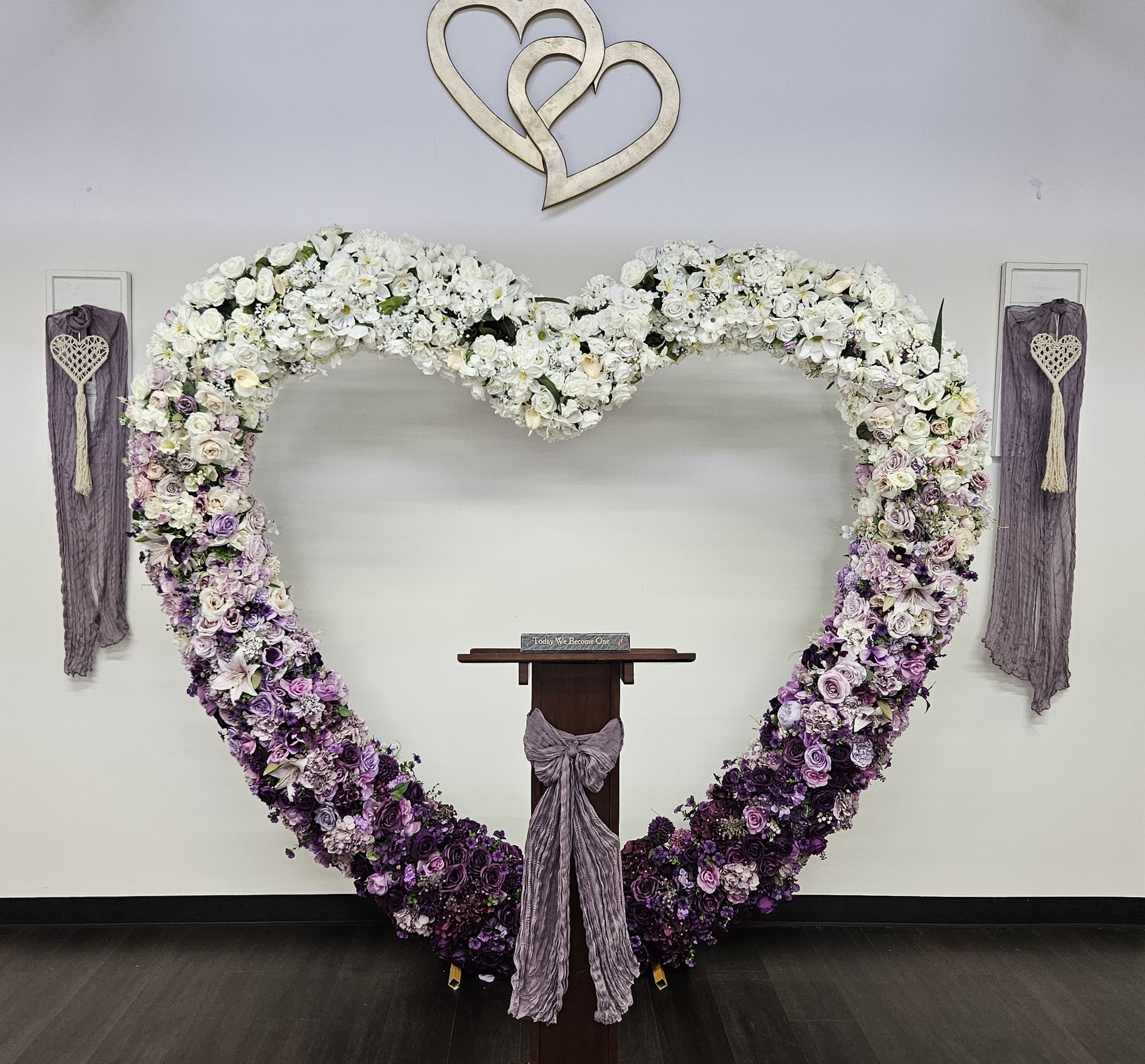 Heart-shaped floral arch in white and purple, over a wooden stand. Two decorative hanging panels on either side.