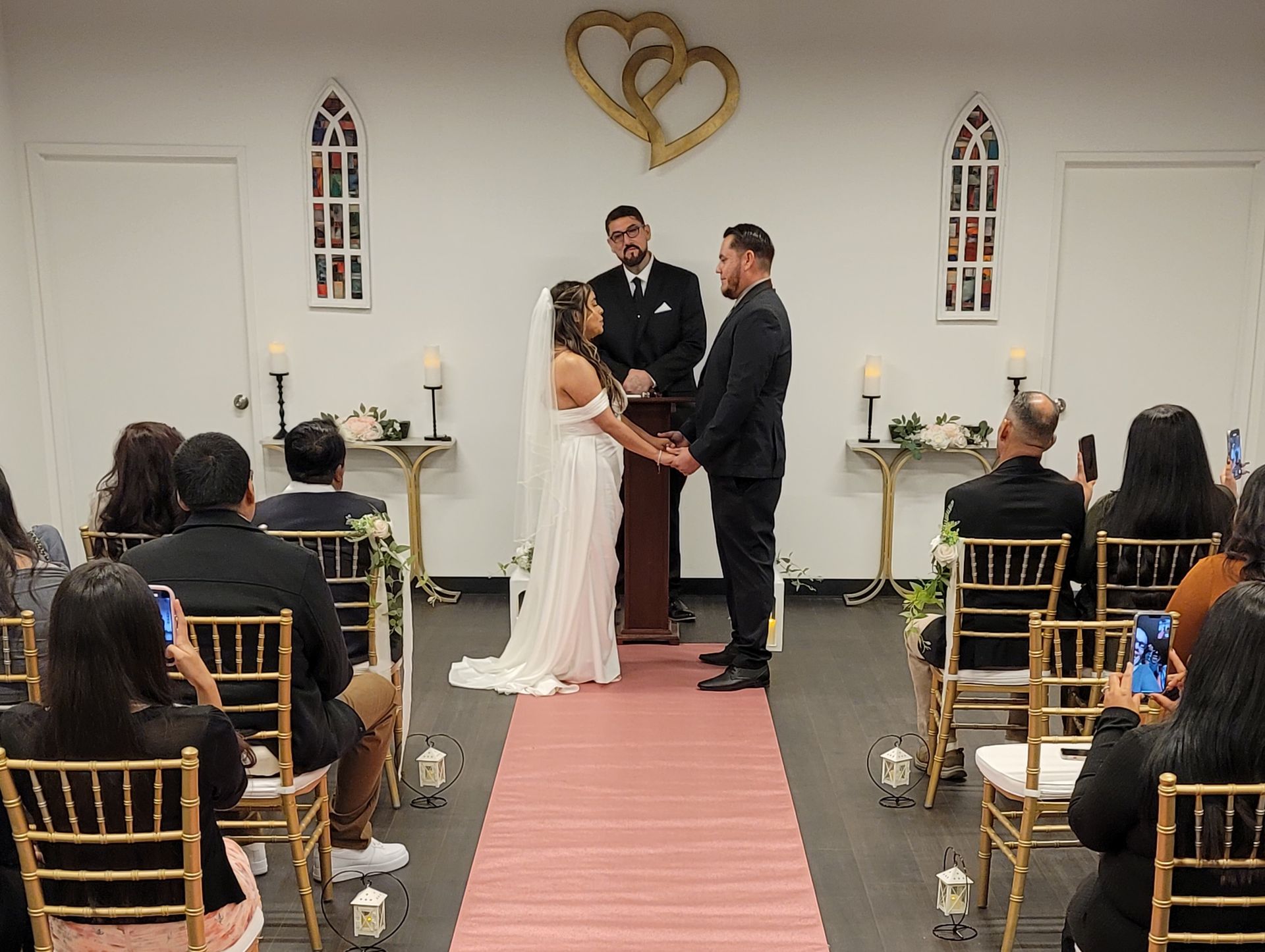 Wedding ceremony: couple holding hands, facing officiant, chapel setting, guests seated, pink aisle.