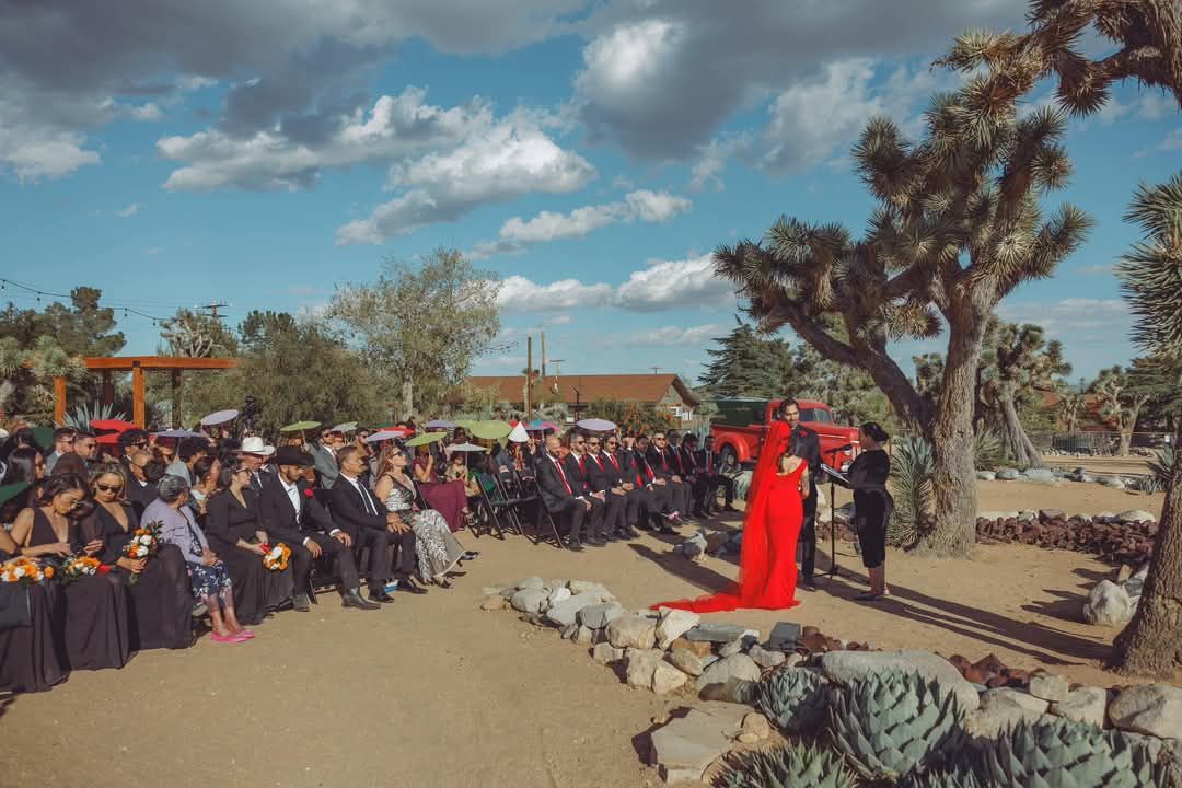 Wedding ceremony in desert: Bride in red, guests seated, officiant, Joshua trees, blue sky.