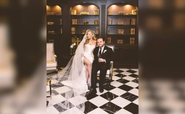 Newlyweds pose indoors: woman in white dress, man in tuxedo, checkered floor, shelves in background.