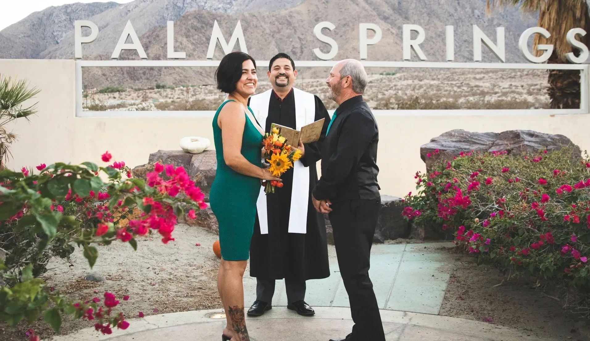 Couple getting married in Palm Springs, California. A smiling officiant, bride in green dress, groom in black suit.