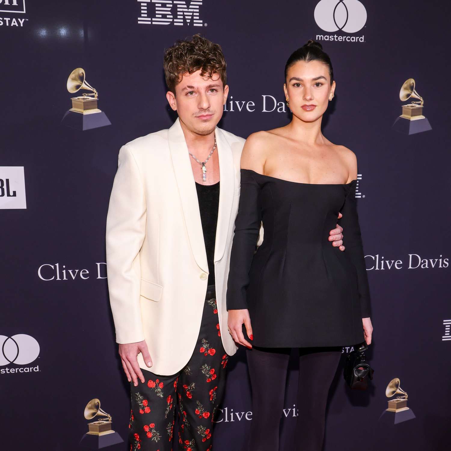 Charlie Puth and Brooke Sansone pose at an event, he in cream blazer and floral pants, she in a black off-shoulder dress.