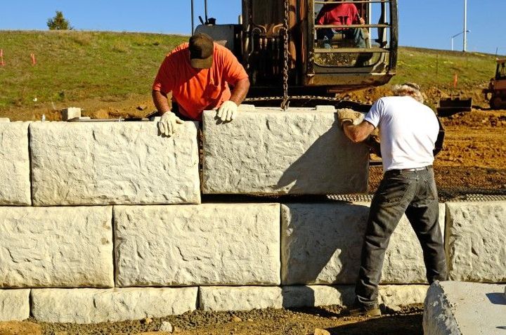 Two construction workers in gloves position large concrete blocks to build a wall.