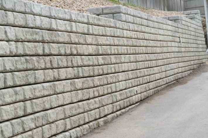 A long wall made of stacked, textured stone blocks runs parallel to a sidewalk. 