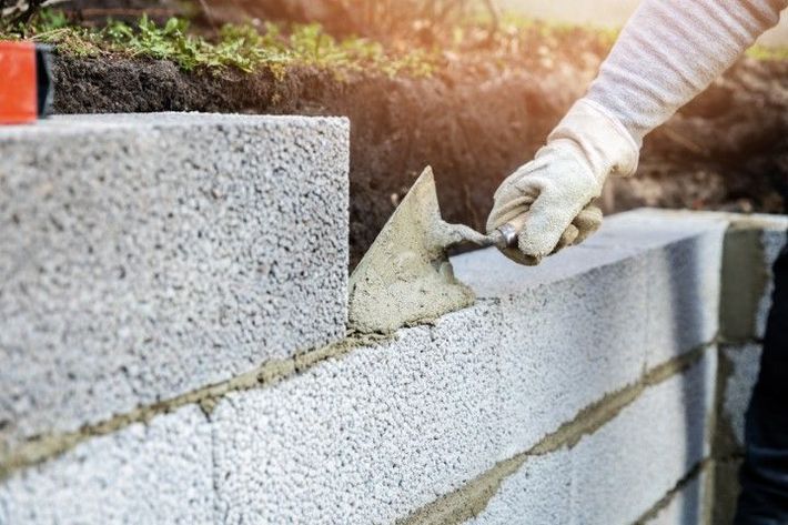 A worker, wearing gloves, uses a trowel to apply mortar between concrete blocks, building a wall.