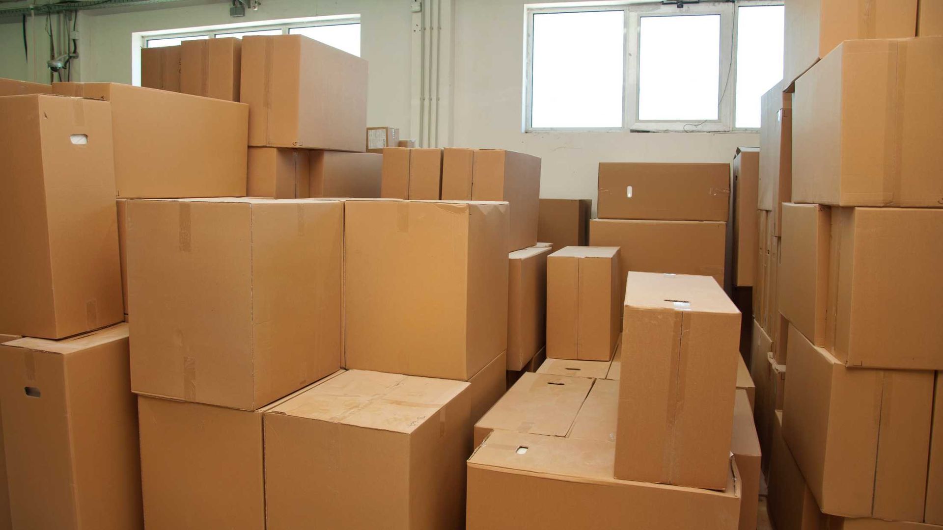 Cardboard boxes stacked in a warehouse near a window, suggesting storage or inventory.