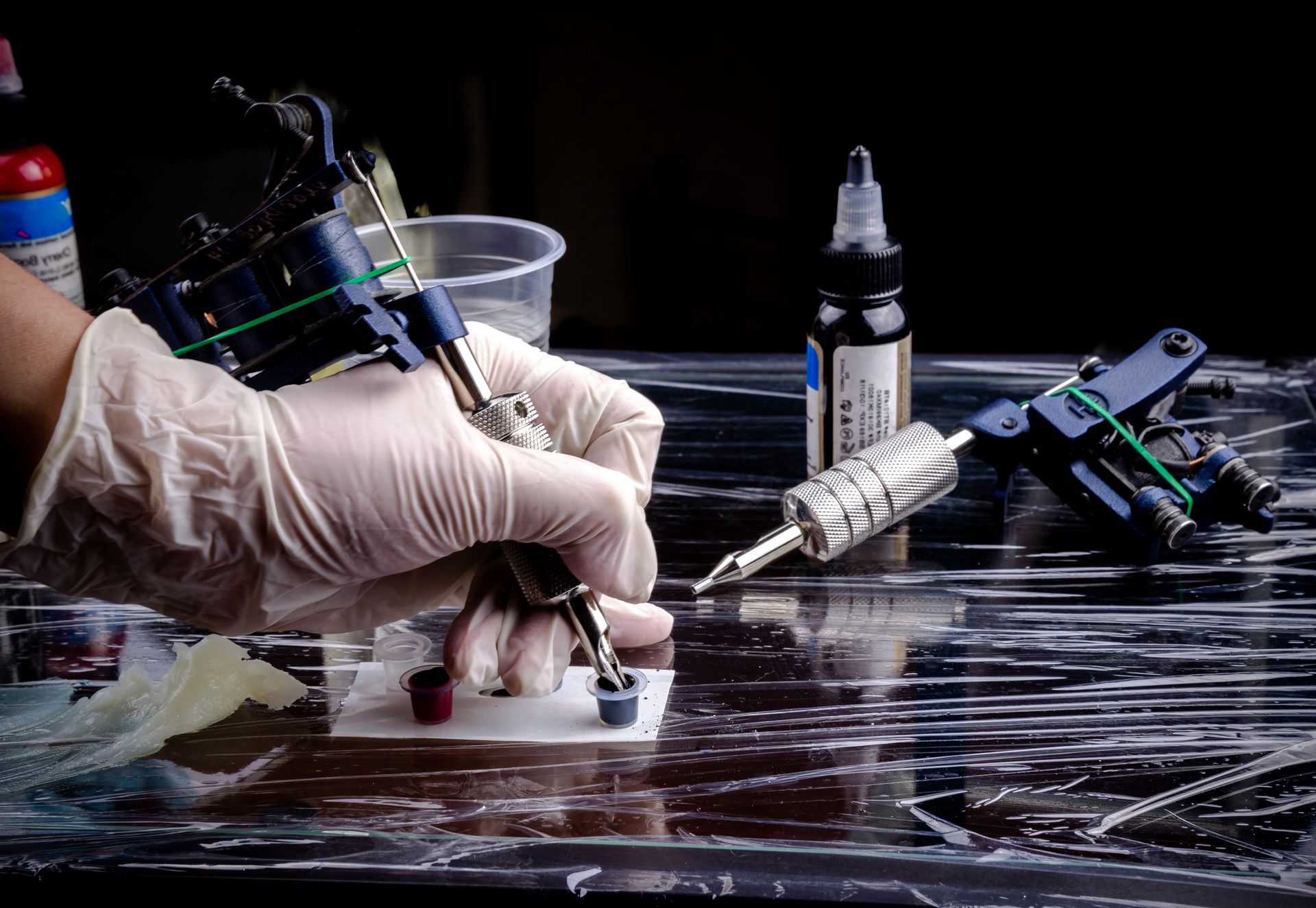 Gloved hands tattooing on skin beside ink bottles and tools on a plastic-covered table