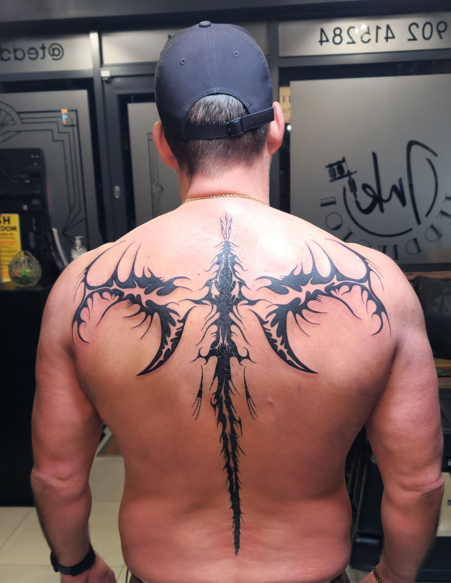 Back view of a shirtless man wearing a cap, showing a large black tribal tattoo across his upper back.