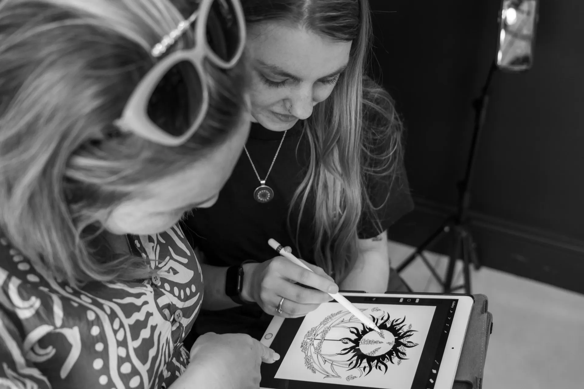 Two people sketching a sun design on a tablet in black and white.