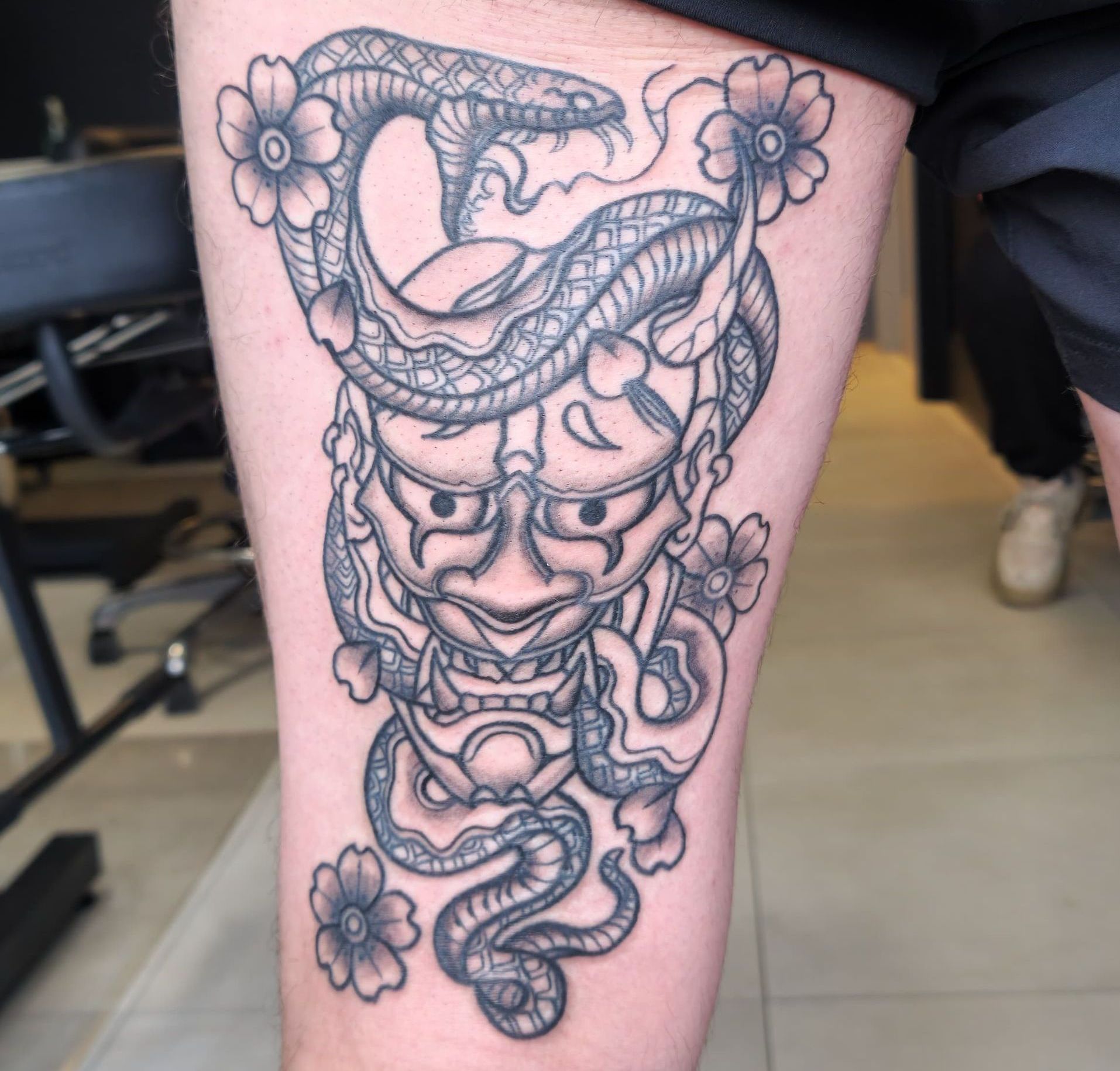 Black linework tattoo of a horned mask with a snake and flowers on a leg
