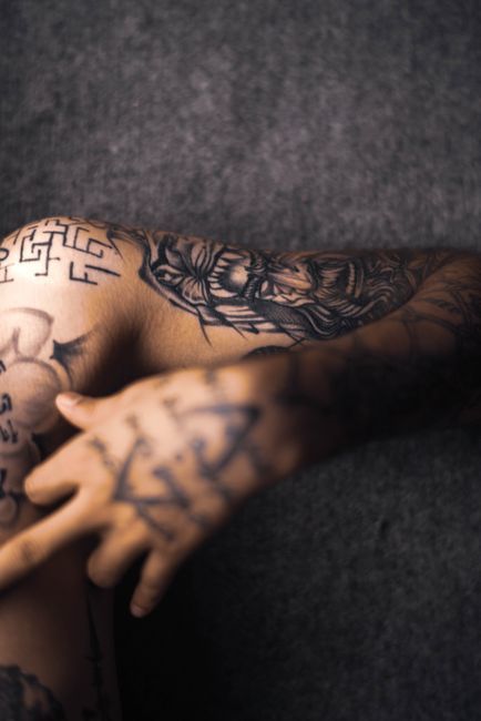 Tattooed arm and hand resting on a dark gray couch, with a blurred black-and-white tattoo design visible.