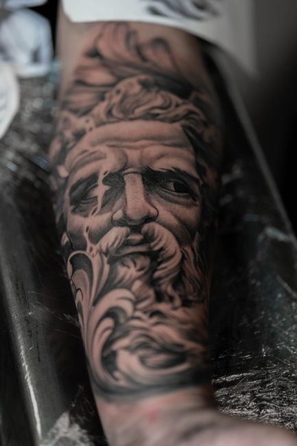 Black-and-gray portrait tattoo on a forearm, showing a detailed face with flowing smoky shading