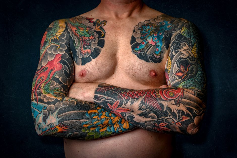 Torso of a shirtless person with colorful full-sleeve and chest tattoos, arms crossed against a dark background